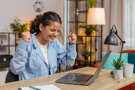 Happy young Indian woman working on laptop netbook shocked by sudden victory, winning, goal achievement, good career news. Excited freelancer girl clenching fists at home office workplace. Lifestyle. - Powered by Adobe