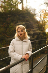 Blonde woman in a white puffer jacket standing on a nature park pathway, bathed in warm backlight...