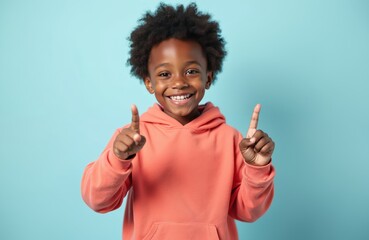 Happy african american boy points up with fingers. Smiling child has positive emotion. Black boy wearing red hoodie, showing something up with index fingers on blue background.