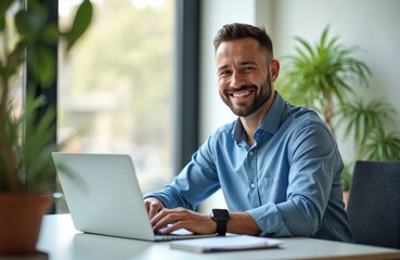 Fototapeta premium Smiling man works on laptop in modern office. Businessman in blue shirt. Happy, successful male entrepreneur typing at desk. Technology, remote work, online business concept.