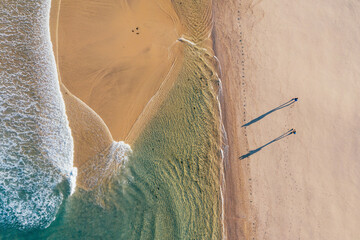 Top-down view of couple walking along sandy beach washed by Atlantic Ocean waves in Portugal casting long shadows in low sunrise light. Shot during the Camino Portuguese —  popular pilgrimage route