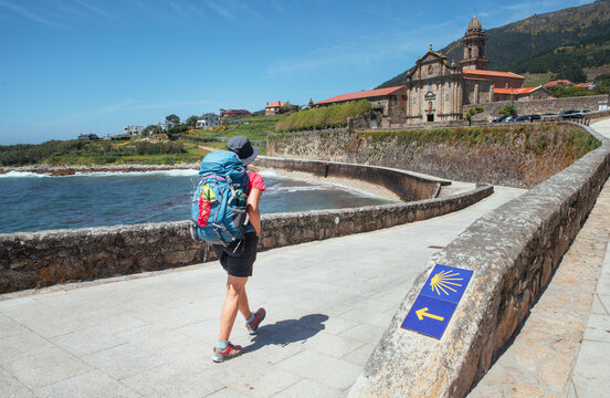 Female pilgrim with backpack walking next yellow arrow and shell directing Camino WAY inspiring solo walk on famous Camino de Santiago, Praia de Oia embankment, Royal Monastery of Santa Mar&iacute;a de Oia