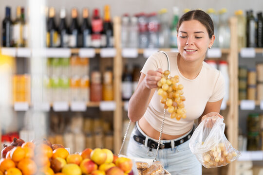 Fototapeta In self-service minimarket, girl customer takes out grape from showcase, examines fruits and buys goods. She picks ripe fruits berries from large box in stores storage
