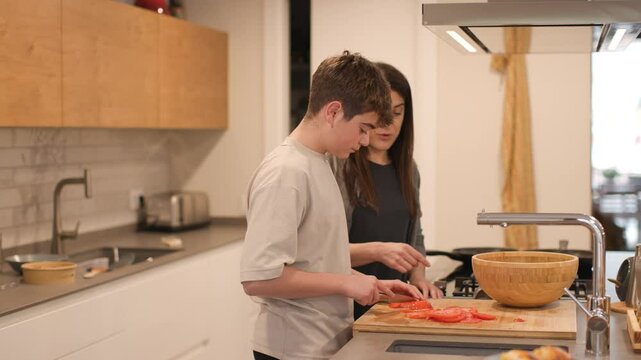 Medium shot of a 14-year-old boy slicing a tomato on a wooden board in a modern, well-lit kitchen. His mother watches and assists nearby. Real family teamwork at home. Horizontal, 4K