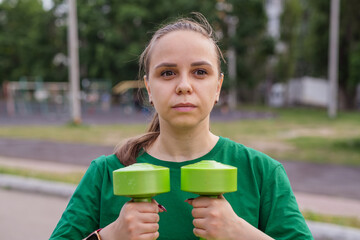A woman in a green t-shirt performs a dumbbell shoulder press exercise during an outdoor workout...