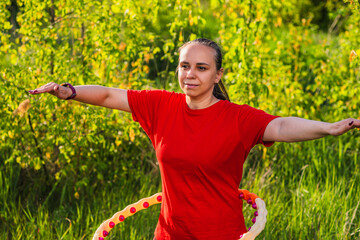 A woman is using a weighted hula hoop while wearing a red shirt and black pants. She is in a green...
