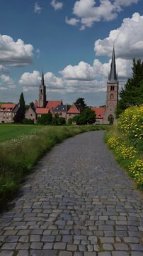 Aerial video view of a serene rural landscape with lush green fields and a quaint village under a sky dotted with fluffy clouds.
