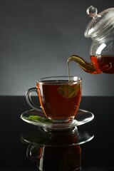 Pouring freshly brewed tea from teapot into cup at dark mirror table, closeup