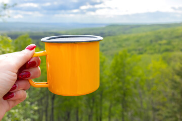 A woman's hiker hand holding a bright yellow metallic cup on the top of a mountain in front of breathtaking view of a forested valley in sun light. Camping and hiking vibes of adventure.