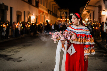 Mujer sahumadora vestida con traje típico tradicional participa en una procesión nocturna durante la Semana Santa en Popayán, Colombia. Sostiene un ramo de flores mientras esparce sahumerio 