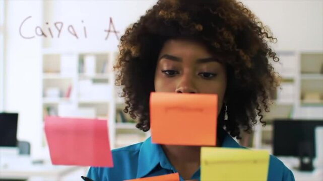A focused Black woman is using colorful sticky notes to organize her thoughts during a brainstorming session in a modern office. The environment is bright and encourages creativity
