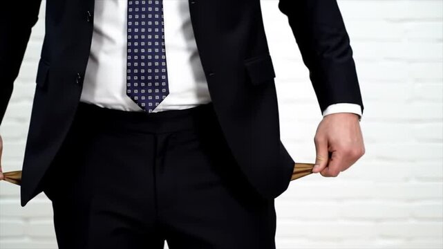 A man in formal attire stands in a contemporary office space, showing his empty pockets as a clear sign of financial struggle. His expression reveals concern and anxiety about money matters