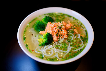 A vibrant bowl of salmon noodle soup, garnished with broccoli florets and sesame seeds, sits on a dark background