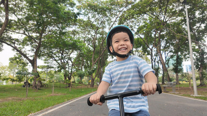 Obraz premium Happy Asian boy riding his balance bike joyfully in a vibrant park filled with trees and greenery, showcasing the joy of outdoor activities.