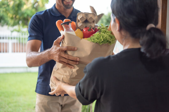 Black male Delivery person hands over a brown paper bag filled with fresh vegetables and fruits to a smiling customer in a residential area.