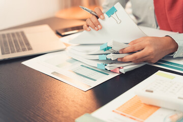 Professional woman checking charts and financial data with calculator and laptop and analyzing and reviewing. Close-up of young woman working with paper documents on desk.