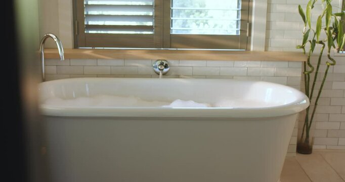 After fill, soapy foam shifting, rippling across freestanding tub water beneath shuttered window