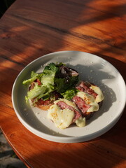 Open-faced sandwich topped with melted cheese, salami, and fresh greens on a rustic plate. The wooden tabletop adds a cozy touch to this inviting and delicious meal setting.