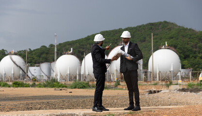 Two businessmen or plant project managers wearing suit and safety hats are holding drawings, discussing, and shaking hands on a business cooperation at Refinery and gas petrochemical plant