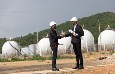 Two businessmen or plant project managers wearing suit and safety hats are holding drawings,...