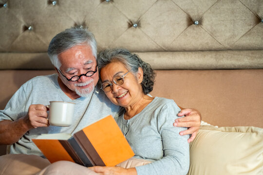 Happy Asian family senior couple relaxing and reading a book together on the bed in bedroom. Elderly husband and wife taking care each other. Retired people healthcare and mental health concept. - Powered by Adobe