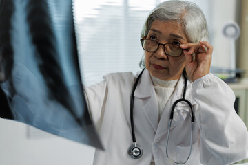 Experienced Asian female doctor in white coat adjusting glasses while carefully examining chest...