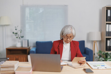 Focused elderly asian businesswoman wearing red jacket and glasses sitting at desk and reviewing financial documents while working remotely from home office