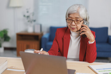 Focused Asian female entrepreneur in red jacket talking on mobile phone and working with laptop...