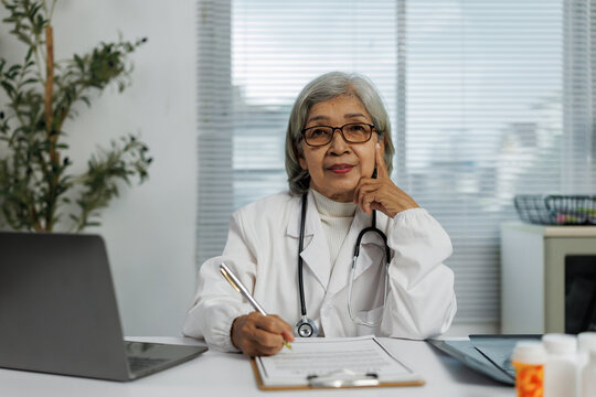 Experienced Asian female doctor wearing a lab coat and stethoscope, diligently writing notes on a clipboard while seated at her desk in a professional medical office setting