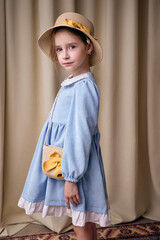 A cute girl with a serious expression poses half-turned. Her hair is tucked under a straw hat. The model is wearing an elegant velvet dress with frills on a beige background