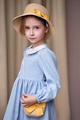 A seven-year-old girl with soft features looks thoughtfully at the camera. She is wearing a set of straw accessories, a woven hat with a yellow ribbon and a shoulder bag. The model in a romantic look 