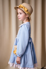 A little girl stands in profile in a light blue dress with white frills at the bottom. On her head she has a straw hat with a yellow ribbon and bow. A light curtain is visible in the background.