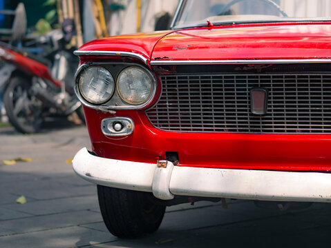 Close-up of a vintage red car with classic round headlights and chrome grille, showcasing aged details and retro styling, parked on an urban street.