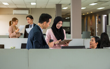 Asian businessman in navy suit discussing tablet content with Muslim colleague in pink hijab at modern office cubicles. Diversity team collaborating in open workspace environment
