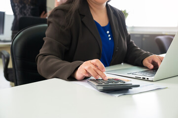 Businesswoman in suit using calculator while working on laptop at office desk. Financial analysis and accounting work in modern corporate environment