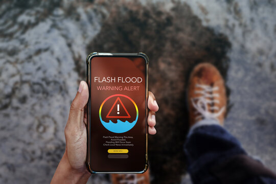 Smartphone displaying a flash flood warning alert screen, held by a person standing near rising floodwaters. Concept for emergency notification, natural disaster preparedness, and weather alerts