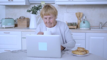 Senior woman sipping coffee while using a laptop in a bright kitchen, savoring her morning routine with delicious breakfast pastries and enjoying a moment of digital connection