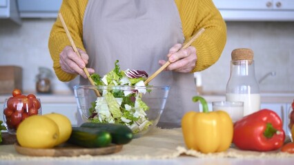 Elderly woman wearing an apron mixing a fresh salad with wooden spoons in a glass bowl in a kitchen...