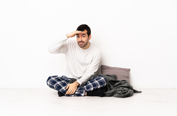 Caucasian man in pajamas sitting on the floor at indoors looking far away with hand to look...