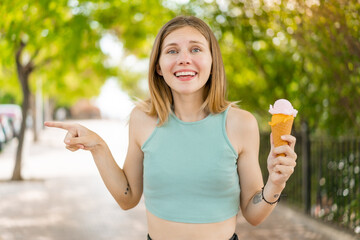 Young blonde pretty woman with a cornet ice cream at outdoors surprised and pointing finger to the...