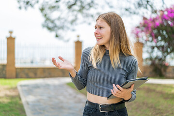 Young blonde woman holding a tablet at outdoors with surprise facial expression