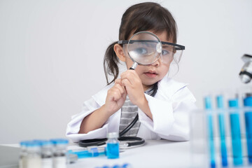 A young child in a lab coat examines a vial with a magnifying glass, showcasing curiosity and interest in science.