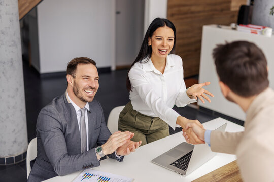 Happy businesswoman standing at corporation and shaking hands with coworker while other colleague is applauding.