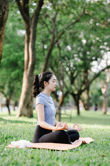 Fototapeta premium Young Asian woman practicing yoga outdoors on a sunny morning in a park, eyes closed, embracing calm, balance, and mindfulness.