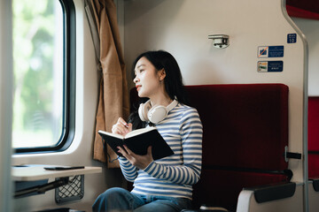 Asian woman sitting at a railroad station with backpack, using mobile phone and laptop, waiting for...