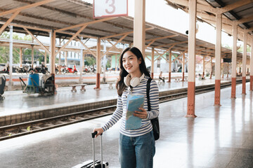 Fototapeta premium Asian woman sitting at a railroad station with backpack, using mobile phone and laptop, waiting for train, enjoying travel lifestyle.