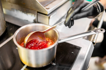 A professional chef stirs a vibrant red tomato sauce in a stainless steel pot using a ladle, wearing a black glove. The scene captures a culinary moment in a kitchen, highlighting food preparation