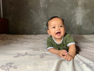 An Asian infant boy lying on his stomach on a bed, smiling joyfully with mouth open. Captured in soft natural light with a neutral wall background in a cozy indoor setting.