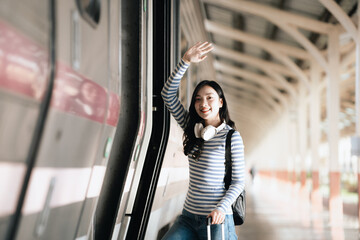 Asian woman sitting at a railroad station with backpack, using mobile phone and laptop, waiting for train, enjoying travel lifestyle.