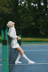 Young female tennis player in stylish sportswear enjoying a moment of relaxation on the court,...